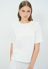 Camiseta de algodón blanca con intrincados bordados de lentejuelas en tono marfil en la parte frontal, escote redondo y mangas cortas. Corte sencillo y recto.