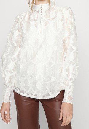 Femme portant un chemisier en dentelle blanche transparente avec un col haut boutonné et des manches longues bouffantes, associé à un pantalon en cuir marron.