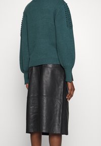 Pull en tricot bleu sarcelle avec manches texturées et ourlet côtelé, associé à une jupe midi en cuir noir élégante avec des détails de coutures.