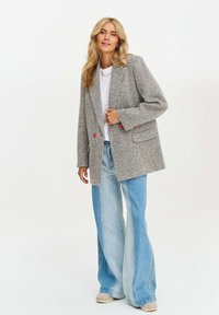 Blazer s houndstooth vzorem v odstínech šedé s růžovými akcenty, s uvolněným střihem, velkými kapsami a hladkým povrchem. Spárován s rozšířenými džíny.