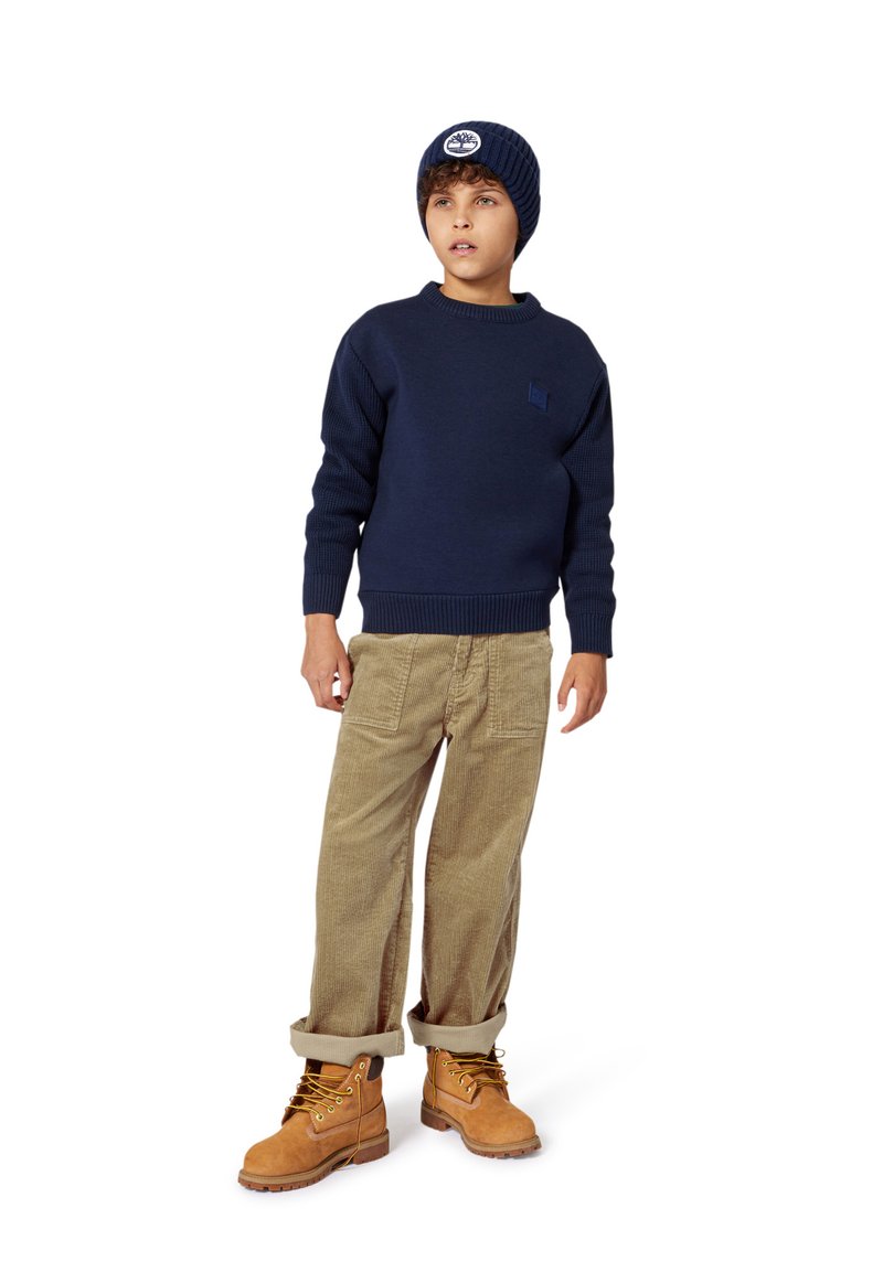 Marine strikkegenser, cordfløyelsbukser i beige og brune støvler. Lue med logo; teksturerte ermer; oppbrettede mansjetter på buksene. Uformell antrekk.