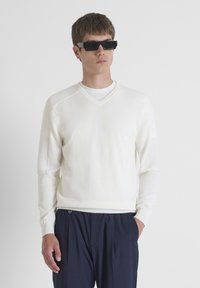 Maglione bianco a collo a V con dettagli testurizzati sulle spalle, maniche lunghe e polsini a costine, abbinato a pantaloni scuri e occhiali da sole neri.