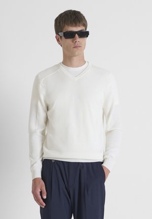 Bijela v-neck pulover s teksturiranim detaljima na ramenima, dugim rukavima i rebrastim manžetama, uparen s tamnim hlačama i crnim naočalama.