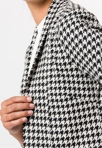 Enfoque cercano de una persona que lleva un blazer de cuadros en blanco y negro sobre una blusa blanca, con una mano ajustando la solapa del blazer.