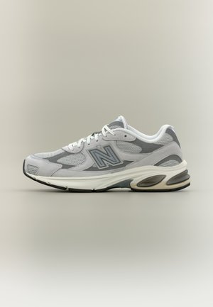 Zapatilla de correr New Balance gris y blanca con paneles de malla y ante, cordones blancos y suela acolchada sobre fondo neutro.