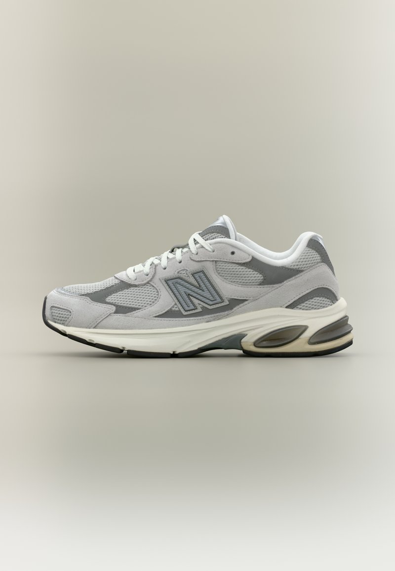 Pantof sport New Balance gri și alb, cu panouri din plasă și piele întoarsă, șireturi albe și talpă amortizată, pe fundal neutru.