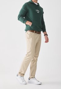 Uomo che indossa un felpa verde, pantaloni beige con cintura marrone, scarpe da ginnastica bianche e un orologio marrone, in piedi contro uno sfondo bianco uniforme.