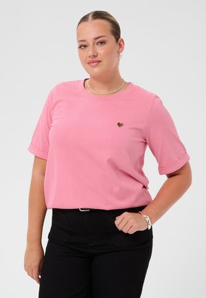 Femme aux cheveux châtain clair porte un t-shirt rose à manches courtes avec un petit détail en forme de cœur et un pantalon noir, posant devant un fond uni.