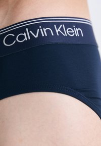 Nahaufnahme einer Person, die dunkelblaue Calvin Klein Unterwäsche mit einem gebrandeten Bund und sichtbaren Nähten trägt.