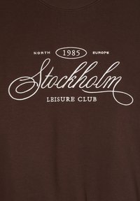 Felpa marrone con collo a girocollo e stampa bianca: "North 1985 Europe Stockholm Leisure Club." Tessuto liscio, design casual.