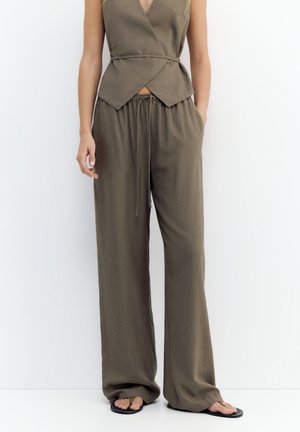 Mouwloze olijfgroene overslagtop gecombineerd met losse, hoge taille broek met trekkoord en zwarte slippers, gedragen door een staande persoon.