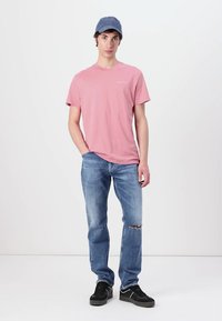 Rózsaszín pamut póló, kék sapka, világoskék, megviselt farmernadrág és fekete sneaker. A modell semleges testtartásban áll fehér háttér előtt.