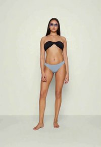 Mulher em pé descalça contra um fundo simples, usando um top de bikini preto torcido, parte de baixo de bikini azul claro e óculos de sol.