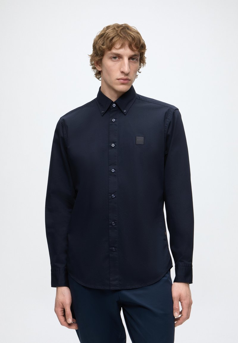 Jeune homme aux cheveux bouclés portant une chemise bleu marine foncé à manches longues boutonnée et un pantalon assorti, debout devant un fond clair uni.