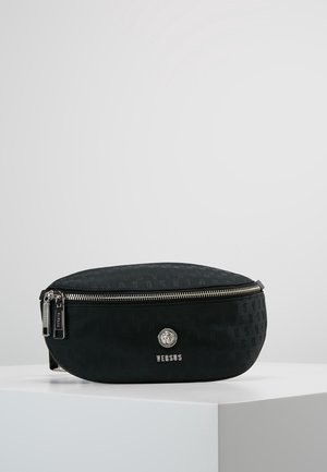 Zwarte Versus riemtas met zilveren rits, reliëflogo patroon en leeuwenkop embleem op een wit voetstuk tegen een grijze achtergrond.