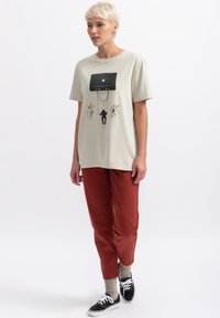 Beige katoenen t-shirt met een zwarte grafische print en kettingen; gedragen met tapse rode broek en zwarte sneakers met witte zolen en veters.