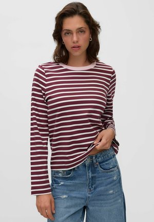 Jeune femme aux cheveux courts et bruns porte un t-shirt à manches longues rayé bordeaux et blanc et un jean bleu délavé, posant devant un fond uni.