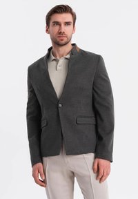 Blazer gris de corte slim, con dos bolsillos frontales y cierre de un botón. Presenta un cuello de muesca y un acento beige en el hombro.
