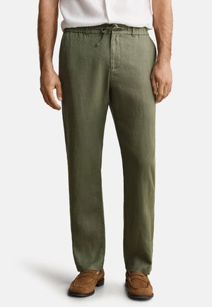 Bărbat purtând pantaloni de in verde măsliniu cu șnur în talie și mocasini maro din piele întoarsă, cămașă albă băgată în pantaloni, stând pe fundal alb.