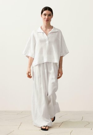 Vrouw loopt naar voren, gekleed in een loszittend wit T-shirt met korte mouwen en wijde witte broek, gecombineerd met zwarte sandalen, tegen een neutrale achtergrond.