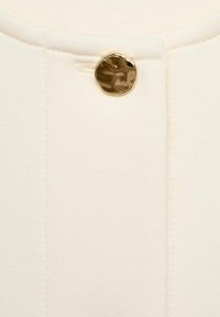 Tessuto di abbigliamento color crema con un bottone rotondo e lucido dorato sulla parte anteriore, arricchito da cuciture verticali per un tocco di dettaglio in più.