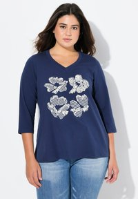 Mörkblå t-shirt med 3/4 ärmar och V-ringning. Framsidan visar en grafik av blommönster i vitt med varierande mönster och strukturer.