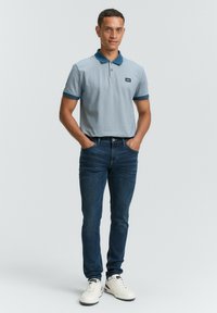 Lichtgrijze polo met een donkerblauwe kraag en manchet, gecombineerd met donkere jeans en witte sneakers. Model staat met de handen in de zakken.