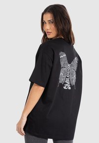 Ung kvinde, der kigger over skulderen, iført en sort oversized t-shirt med et hvidt fingeraftryk "X" design og "SMILODOX" tekst på ryggen.