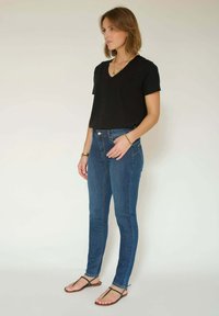 Femme portant un chemisier à col en V noir, un jean skinny bleu et des sandales noires, debout contre un fond clair uni, de profil.