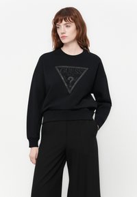 Mujer con cabello rojo vistiendo una sudadera negra con un logo triangular de "GUESS U.S.A." y pantalones anchos negros, de pie contra un fondo blanco.