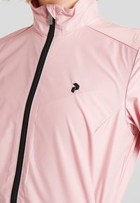 Hellrosa Windbreaker-Jacke aus glattem, leichtem Stoff. Verfügt über einen hohen Kragen und einen schwarzen Reißverschluss mit kleinem schwarzem Logo auf der Brust.