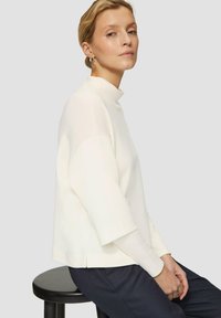 White, high-neck pullover with wide sleeves, featuring a soft texture. Paired with dark patterned trousers. Model seated on a black stool.
