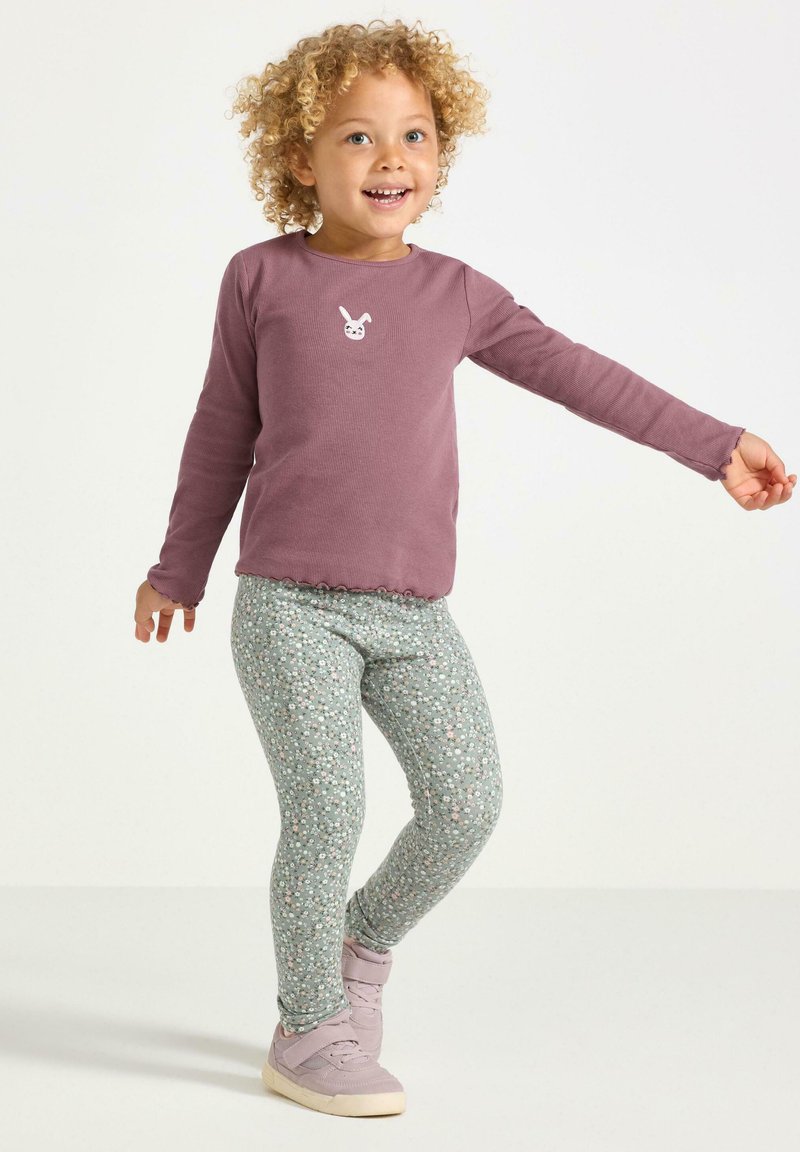 Långärmad skjorta i mauve med vågig nederkant och kaninmotiv, matchad med blommönstrade gröna leggings och ljuslila sneakers.
