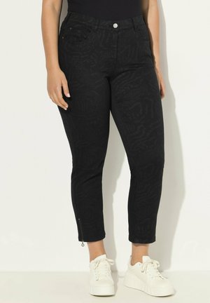Jean en denim noir avec imprimé animal texturé, design ajusté, zips aux chevilles et poches avant. Associé à des baskets blanches.