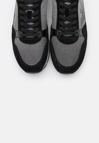 Pari kengät, joissa on mustaa ja hopeaa kimaltavaa kankaita sekä mustat nauhat, kuvattuna ylhäältä valkoista taustaa vasten.