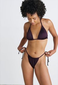 Vijoličen bikini, ki vključuje triangel zgornji del in nastavljive trakove na dnu, izdelan iz gladkega materiala z minimalnim dizajnom in brez vzorcev.