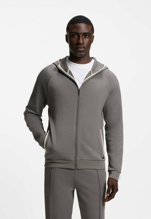 Uomo con felpa grigia con zip e pantaloni abbinati, con le mani nelle tasche, in piedi davanti a uno sfondo bianco uniforme.