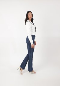 Suéter blanco con detalles negros, jeans acampanados azules y sandalias de tacón blancas destalonadas, todo sobre un fondo liso.