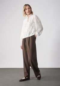 Blusa blanca con estampado floral, de cuello alto y cordón, combinada con pantalones marrones de corte suelto y bailarinas negras.
