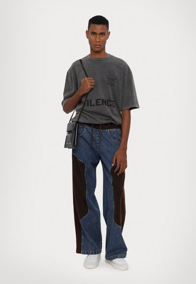 Graues, übergroßes T-Shirt mit "SILENCE"-Druck, kombiniert mit Jeans aus Denim mit braunen Stoffpaneelen und lockerer Passform. Schwarze Umhängetasche. Weiße Schuhe.