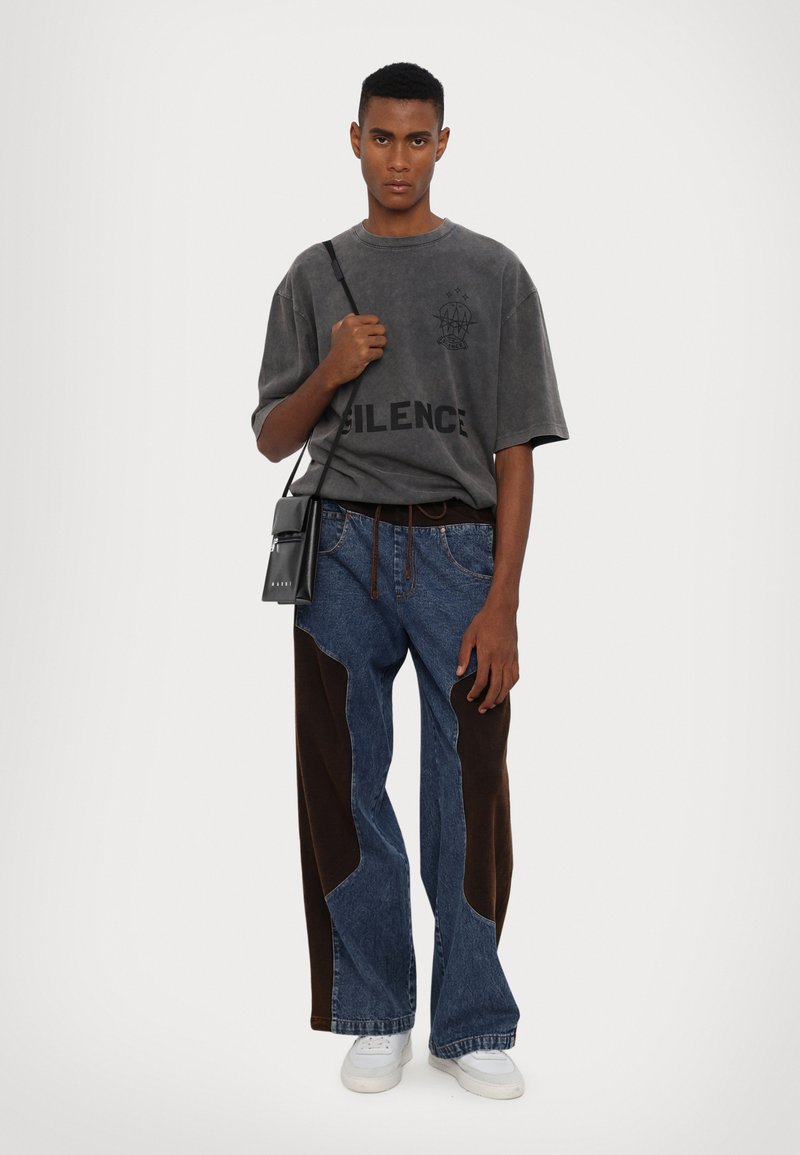 T-shirt grise oversize avec impression "SILENCE", associée à un jean en denim avec des panneaux en tissu marron et une coupe ample. Sac en bandoulière noir. Chaussures blanches.