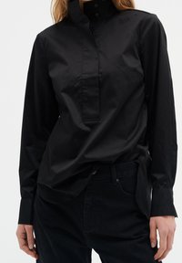 Schwarzes Langarmshirt mit hohem Kragen, ausgestattet mit einer Vorderleiste, aus leichtem Stoff, kombiniert mit dunklen Jeans.