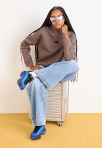 Young woman with long braids wearing reflective sunglasses, brown sweatshirt, wide-leg jeans, and blue sneakers sitting on a metal box.