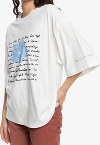 Bílý oversize tričko vyrobené z bavlny, s modrou květinovou grafikou a ručně psaným textem. Zelený text na rukávu říká: "Vložte do toho své srdce."