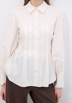 Femme portant un chemisier crème à manches longues, boutonné, avec des plis verticaux, et un pantalon marron, debout devant un fond blanc.