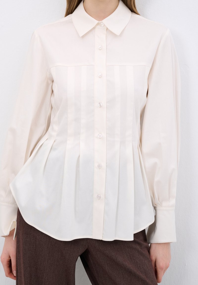 Femme portant un chemisier crème à manches longues, boutonné, avec des plis verticaux, et un pantalon marron, debout devant un fond blanc.