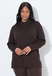 Femme avec de longs cheveux noirs portant un pull à manches longues marron foncé et un pantalon assorti, se tenant devant un fond clair.