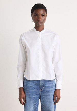 Camicia bianca con colletto, chiusura con bottoni, maniche lunghe e orlo corto. Indossata con jeans in denim blu chiaro. Texture del tessuto liscia.