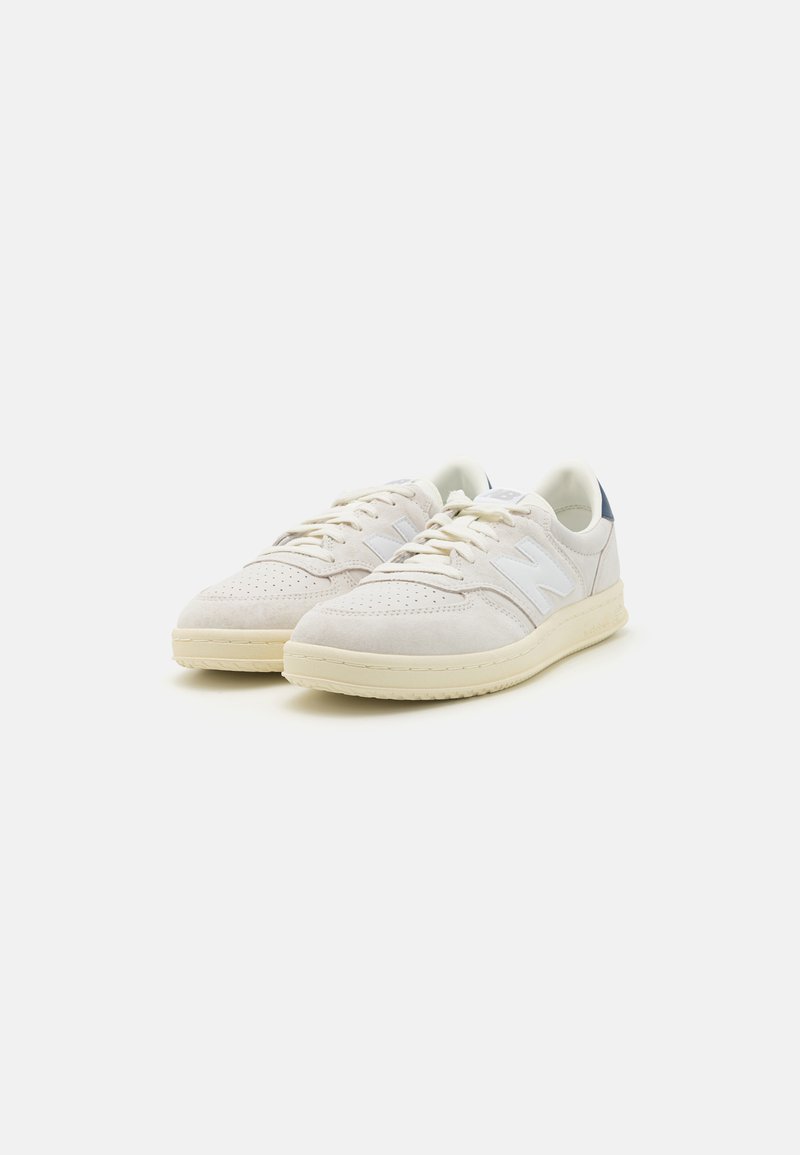 Zapatillas de ante blancas con caja de puntera perforada, cordones de color beige claro y un acento en el talón de color marino; diseño inspirado en lo retro y suela de goma texturizada.