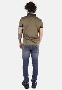 Polo verde oliva con colletto blu navy, indossato con jeans blu scuro. Presenta maniche corte e un design aderente. Tatuaggio visibile sul braccio.